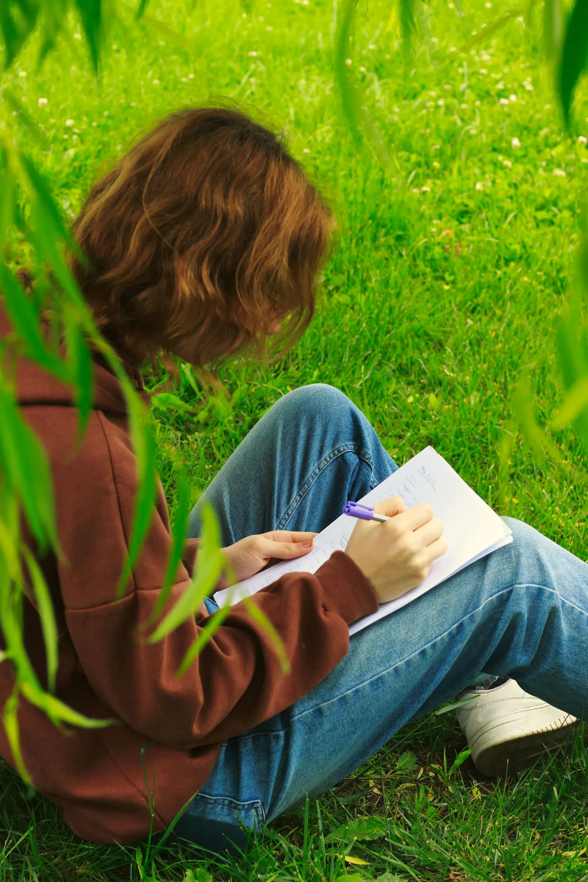 Eine Hand schreibt mit einem Stift in ein Heft, das im Gras liegt.